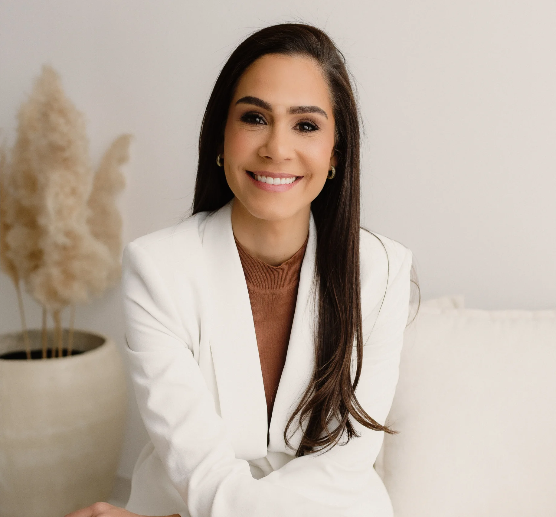Dra. Aline Rezende, cirurgiã plástica, sorrindo com blazer branco sentada em um sofá claro
